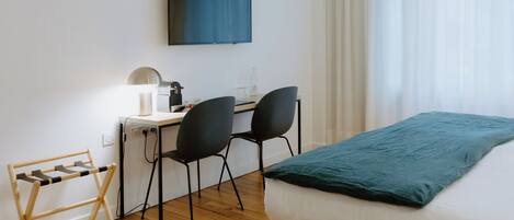 Une chambre d’hôtel comprenant un lit, un bureau avec une chaise, un téléviseur fixé au mur et une fenêtre avec des rideaux légers.