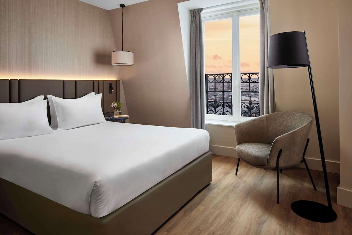 Een hotelkamer met een groot bed, een stoel, een vloerlamp en een raam met uitzicht op de stad.