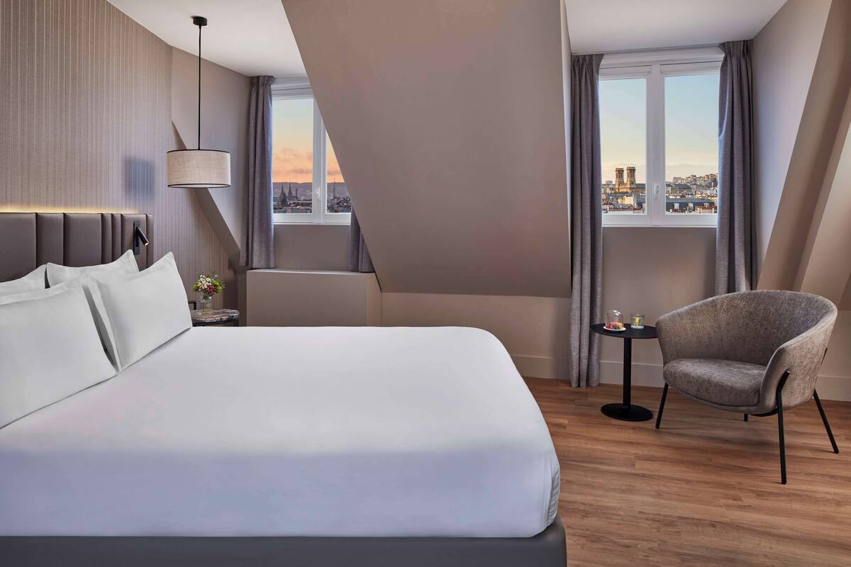 Een moderne hotelkamer met een groot bed, een stoel, een klein tafeltje en uitzicht op de stad door het raam.