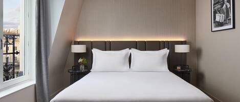 Una habitación de hotel moderna con una cama grande, dos mesitas de noche con lámparas, vistas a la ciudad a través de una ventana y un cuadro en la pared.