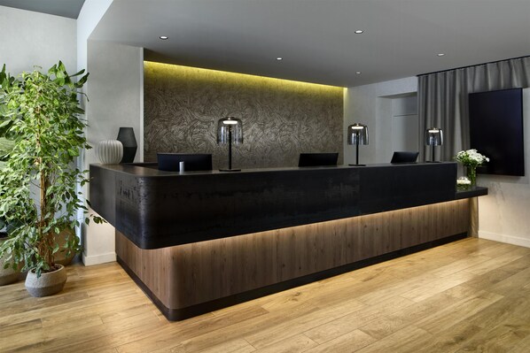 A modern reception desk with a dark countertop, wooden base, and two lamps. A large plant is on the left, and a flat-screen TV is on the right. - NH Paris Gare De l'Est (Paris)