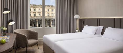 A hotel room with a large bed, a chair, a desk with a lamp, and a view of a building through the window.
