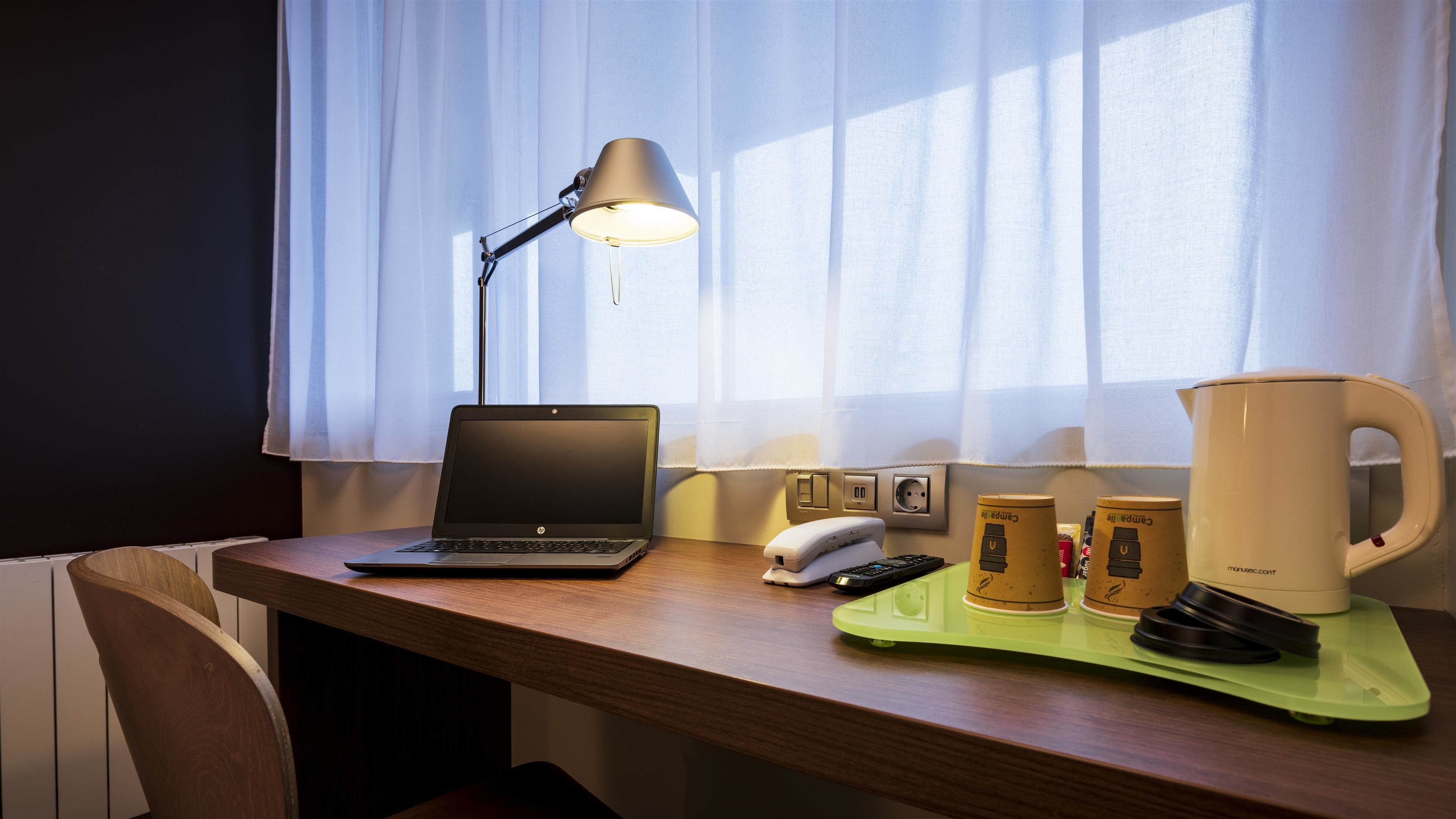 A hotel desk with a laptop, a lamp, a phone, a coffee maker, and a tray with cups.