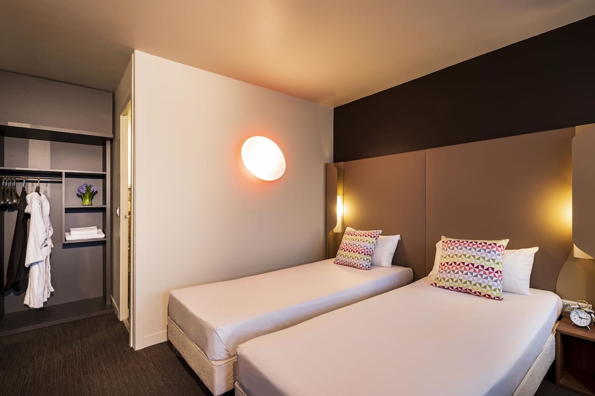 Ein Hotelzimmer mit zwei Einzelbetten, einem Nachttisch mit Lampe, einem weiteren Nachttisch mit einer Uhr, einem Kleiderschrank und einem Badezimmer mit Handtuchhalter.