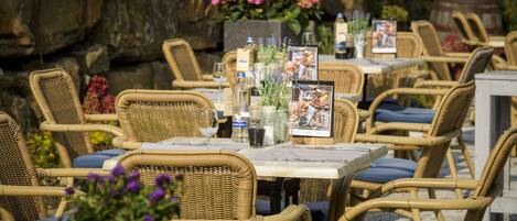 Outdoor wicker tables with blue cushions, arranged in a patio setting with a stone wall and potted plants in the background.