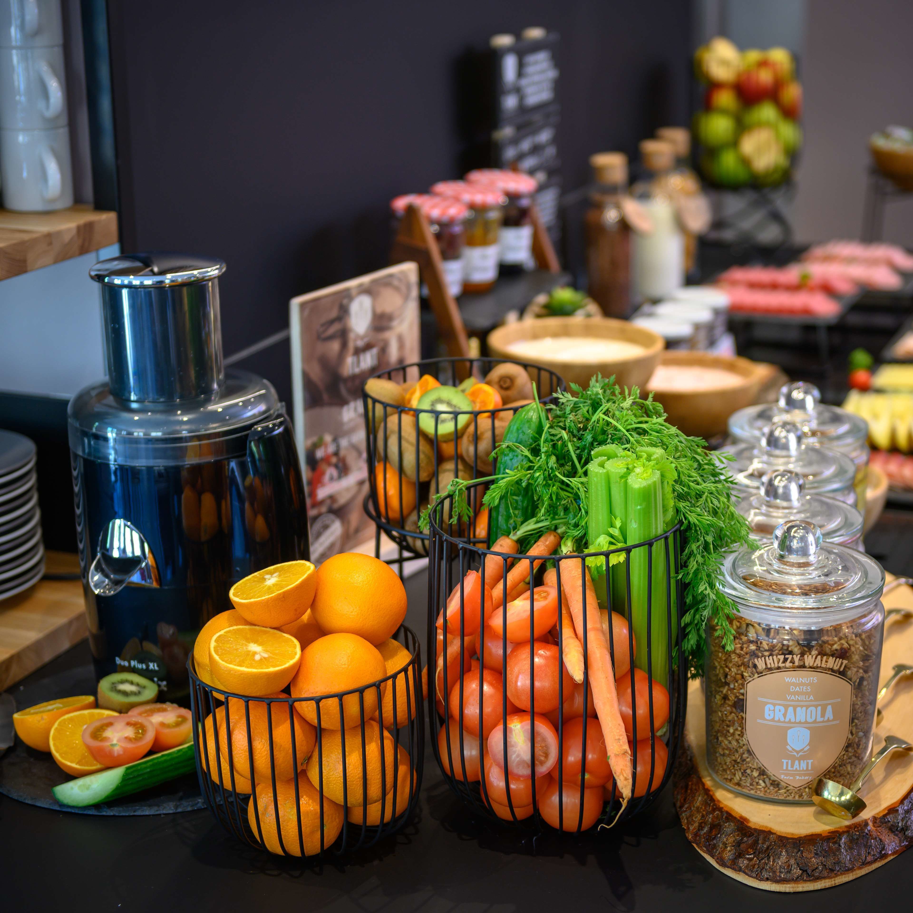 A buffet table with various food items including oranges, tomatoes, carrots, and celery. There are also containers of granola and a water dispenser.