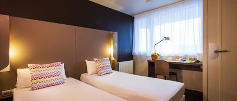 A hotel room with two beds, a desk, a chair, a lamp, and a window with sheer curtains.