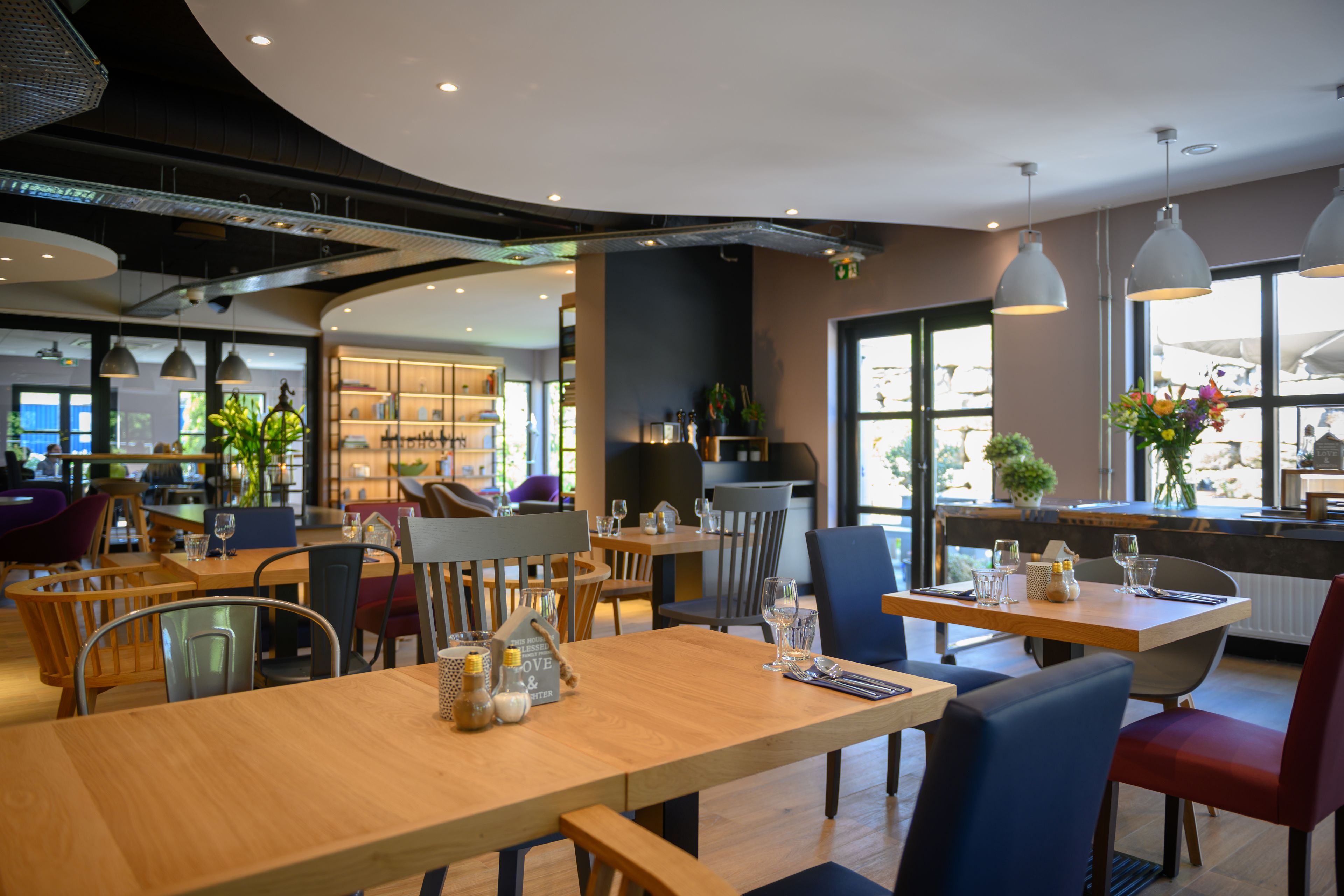 A modern restaurant interior with wooden tables, metal chairs, and pendant lighting.