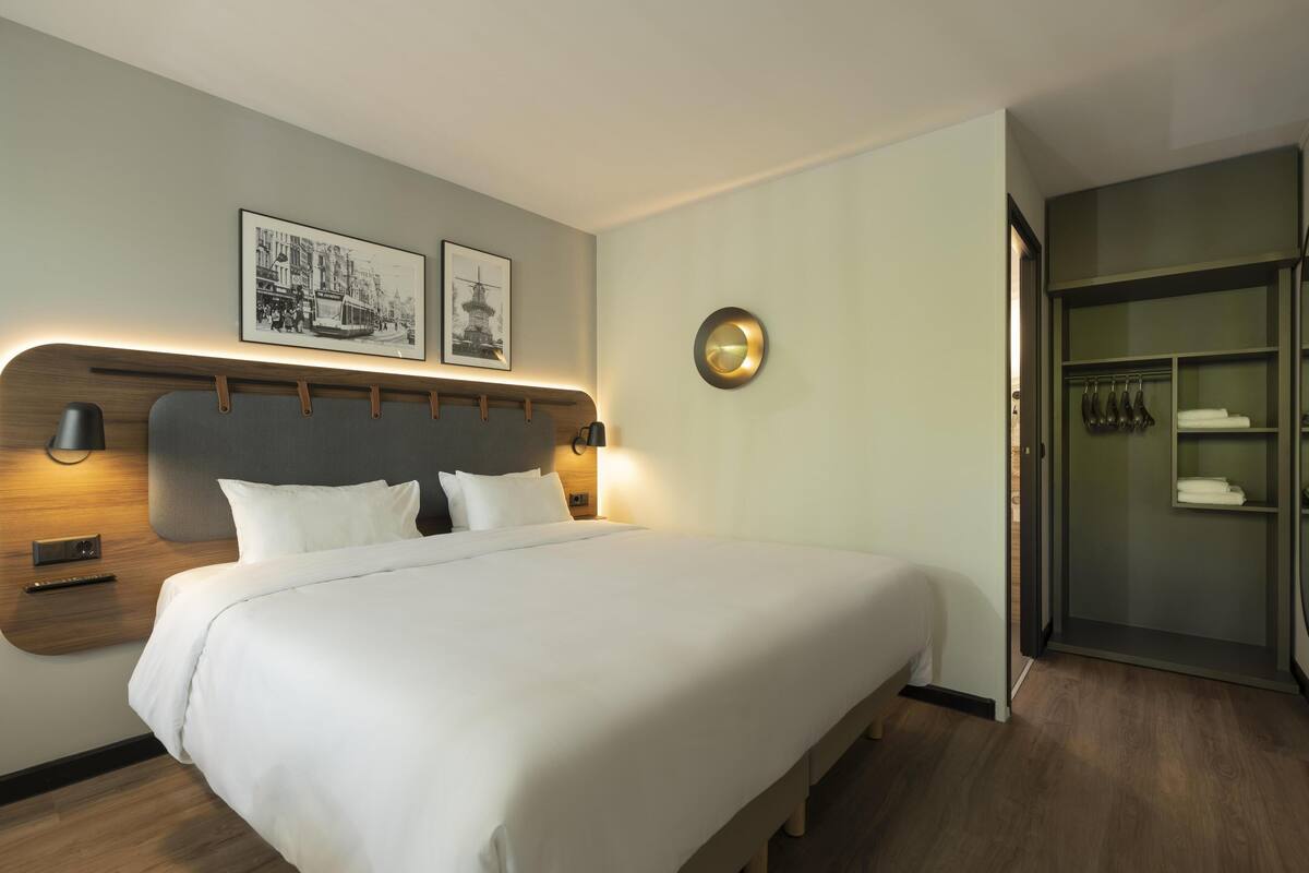 Ein ordentlich eingerichtetes Hotelzimmer mit einem großen Bett, Nachttischlampen, einer Wandleuchte und einem Kleiderschrank mit darin befindlichen Kleidungsstücken.