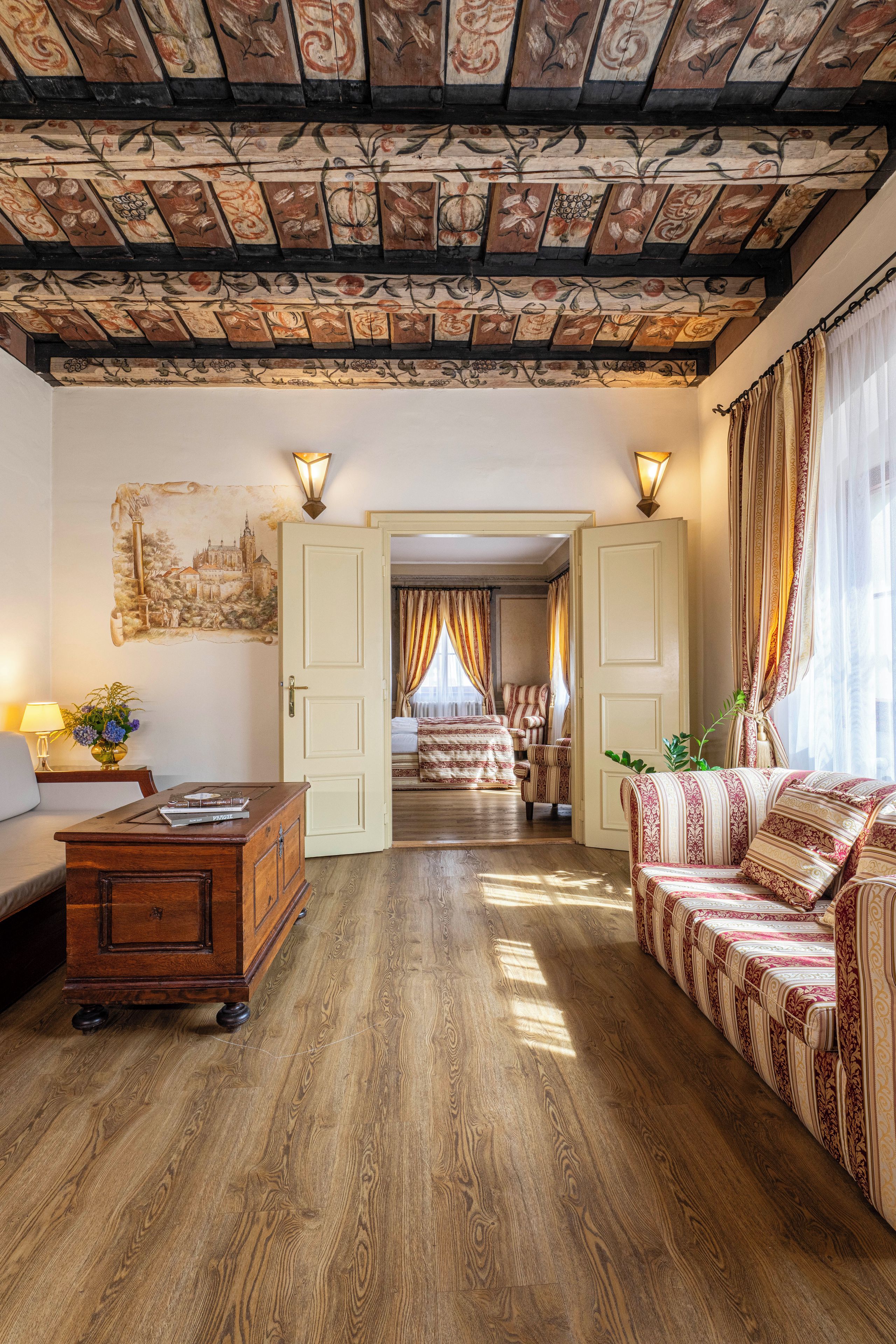A room with a wooden floor, a sofa, a desk, and a bed. There is a painting on the wall and a window with curtains.