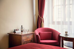 Een hotelkamer met een houten dressoir, een rode stoel met stipjes, een kleine tafel en een raam met dunne gordijnen.