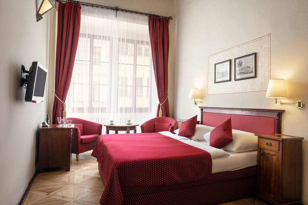 Una habitación de hotel con una cama grande, dos mesitas de noche, un televisor y un ventanal con cortinas rojas.