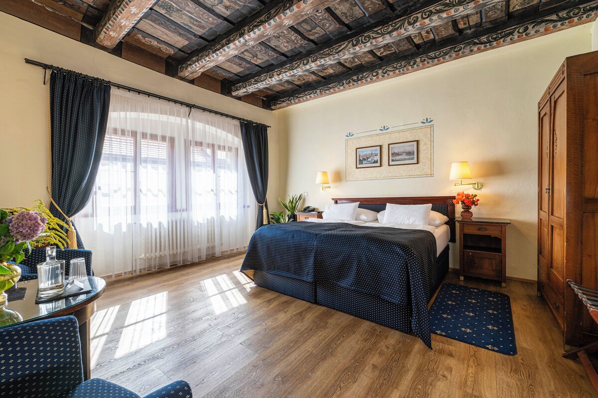Une chambre d’hôtel avec un plafond en bois, un lit recouvert d’une courtepointe bleue, une table de chevet avec une lampe, un bureau agrémenté d’un vase de fleurs et une fenêtre ornée de rideaux légers.