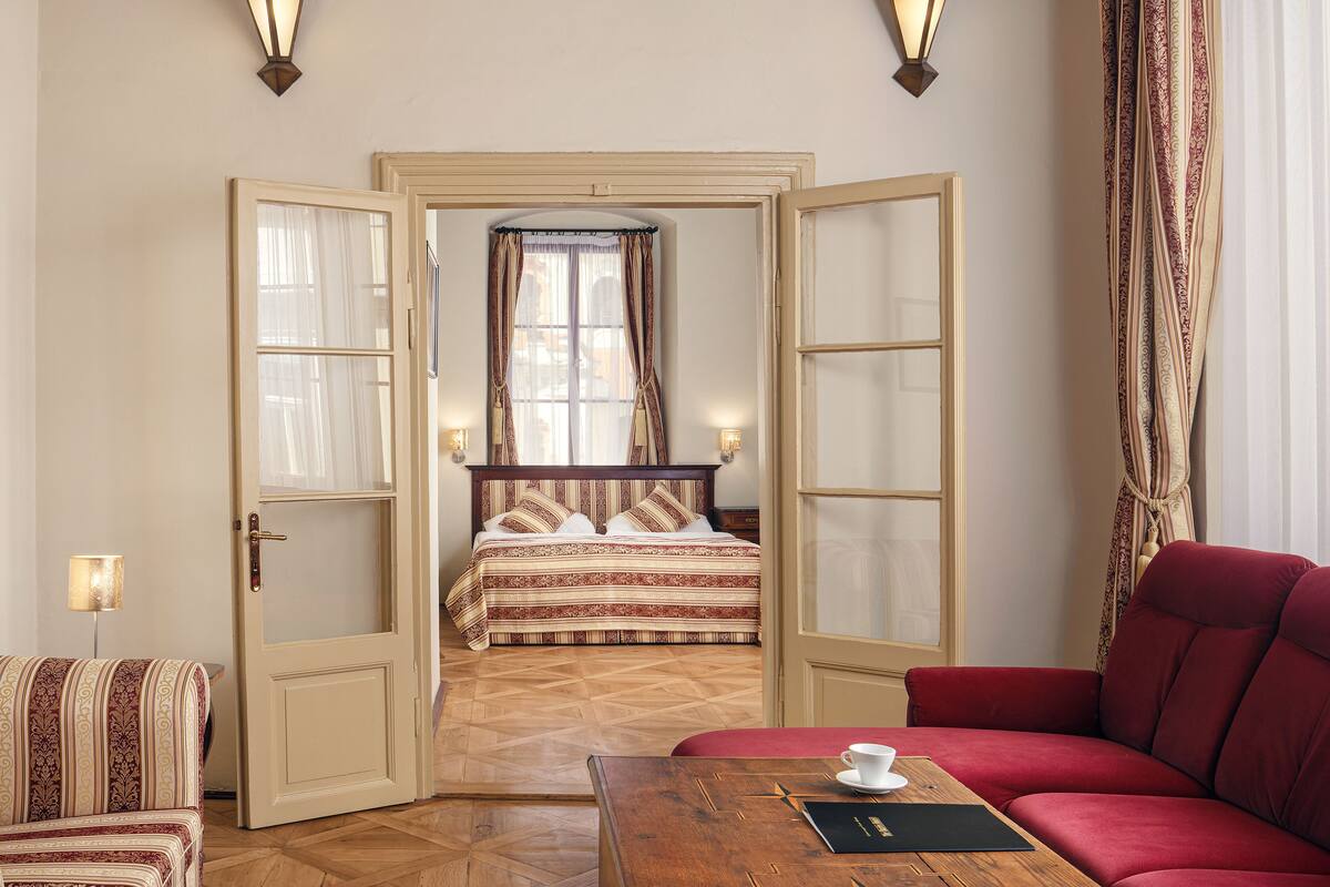 Una habitación de hotel con una cama, un sofá rojo, una mesa de centro de madera y una puerta que da a otra habitación.