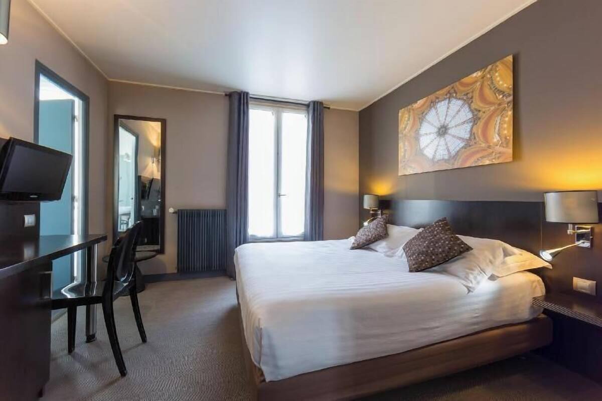 Une chambre d’hôtel avec un lit, un bureau, une chaise, une télévision, un grand tableau et une fenêtre.