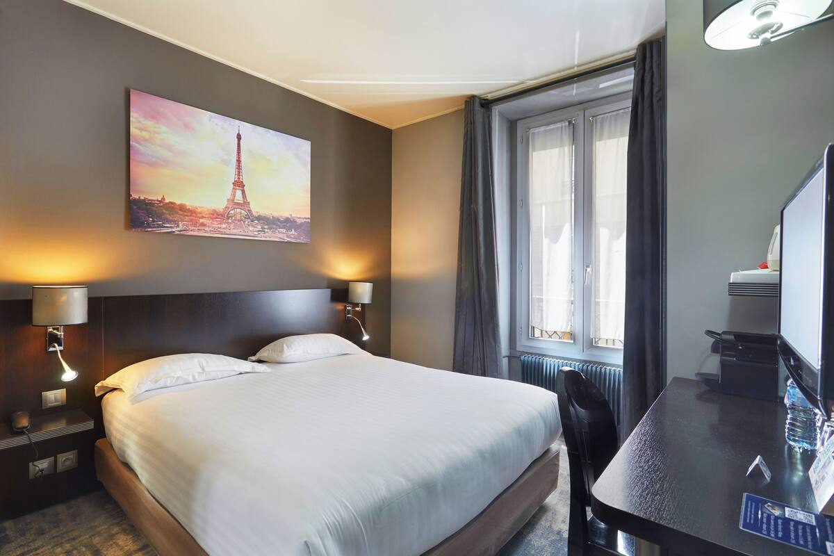 Une chambre d’hôtel avec un grand lit, deux lampes de chevet, un bureau avec une chaise, une fenêtre avec des rideaux et un tableau représentant la Tour Eiffel.