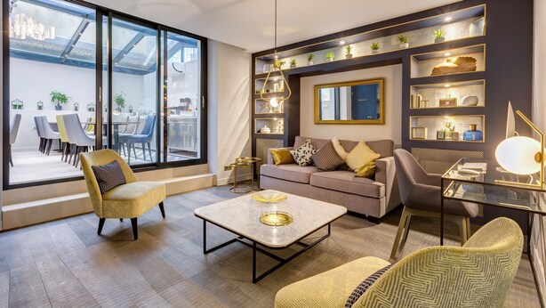 Una sala de estar moderna con sofá, sillones, una mesa de centro y una mesa de vidrio. Hay un ventanal con vistas al exterior y estanterías con objetos decorativos.