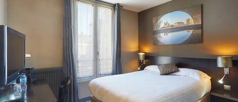 A hotel room with a large bed, a desk with a computer, a TV, and a window with curtains.