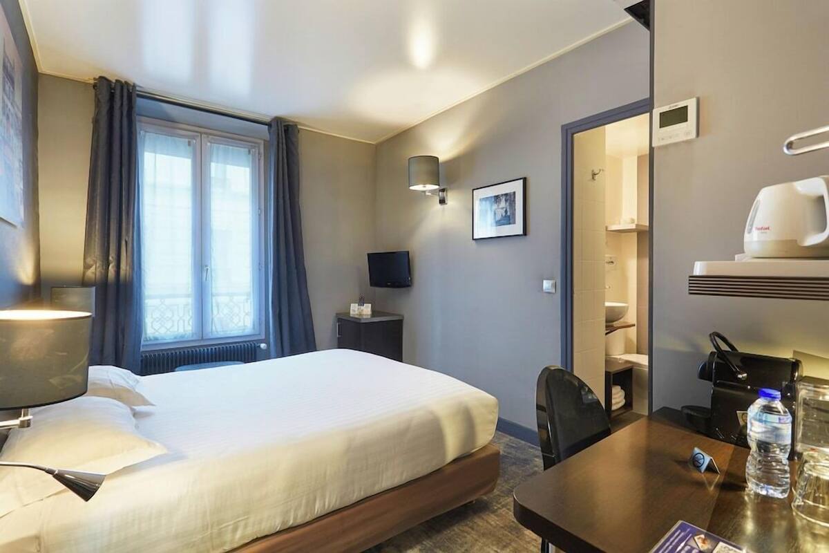Une chambre d’hôtel équipée d’un lit, d’un bureau, d’une chaise, d’une télévision, d’une lampe, d’une bouteille d’eau et d’une machine à café.
