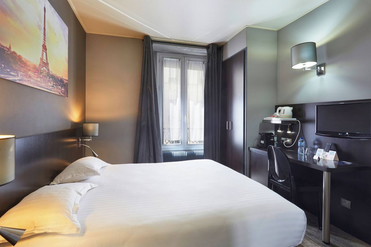 Une chambre d’hôtel avec un grand lit, un bureau avec une chaise, une télévision, une machine à café et une fenêtre avec des rideaux.