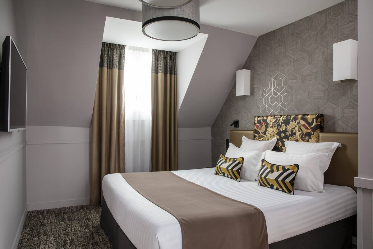 A hotel room with a large bed, a flat-screen TV, a window with curtains, and a geometric patterned wall.