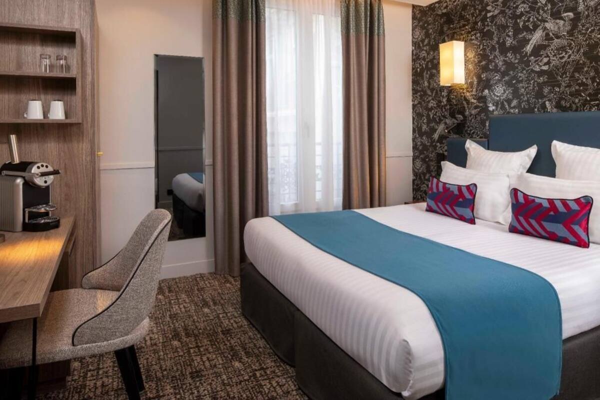 A hotel room with a bed, a desk, a chair, a mirror, and a wall-mounted lamp.