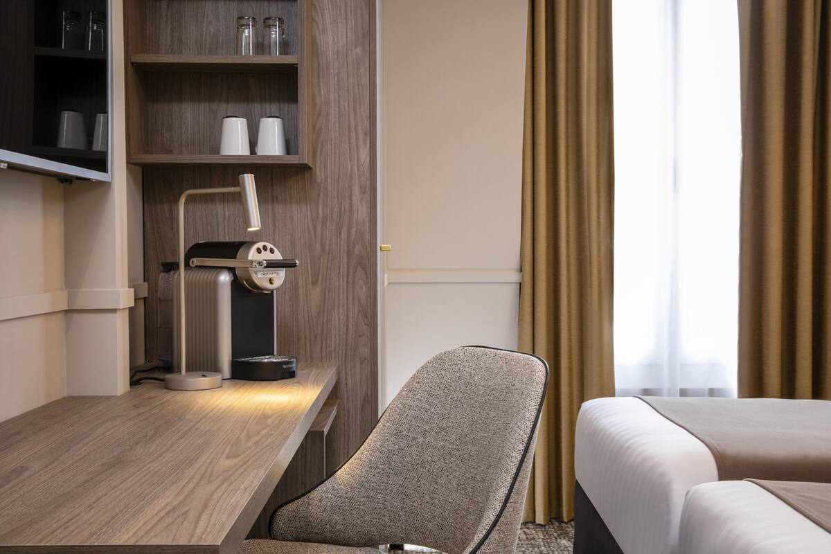 A hotel room with a wooden desk, a lamp, a chair, a bed, and a shelf with cups and a coffee maker.