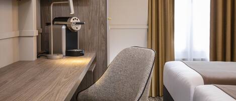 Habitación de hotel con un escritorio de madera, una lámpara, una silla, una cama y una estantería con tazas y cafetera.