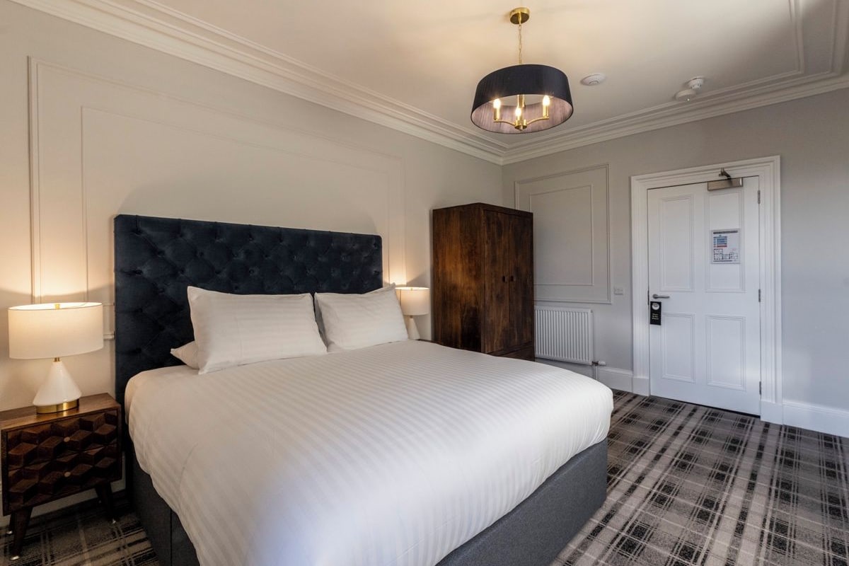 A hotel room with a large bed, a dark headboard, a wooden wardrobe, a white door, and a plaid-patterned carpet.