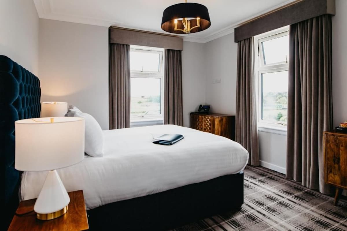 A hotel room with a large bed, a bedside table with a lamp, a small wooden side table, and a view through the window.