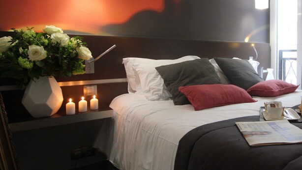 Une chambre d’hôtel avec un lit, du linge de lit blanc, un vase avec des fleurs blanches, trois bougies allumées et un magazine sur la table de chevet.