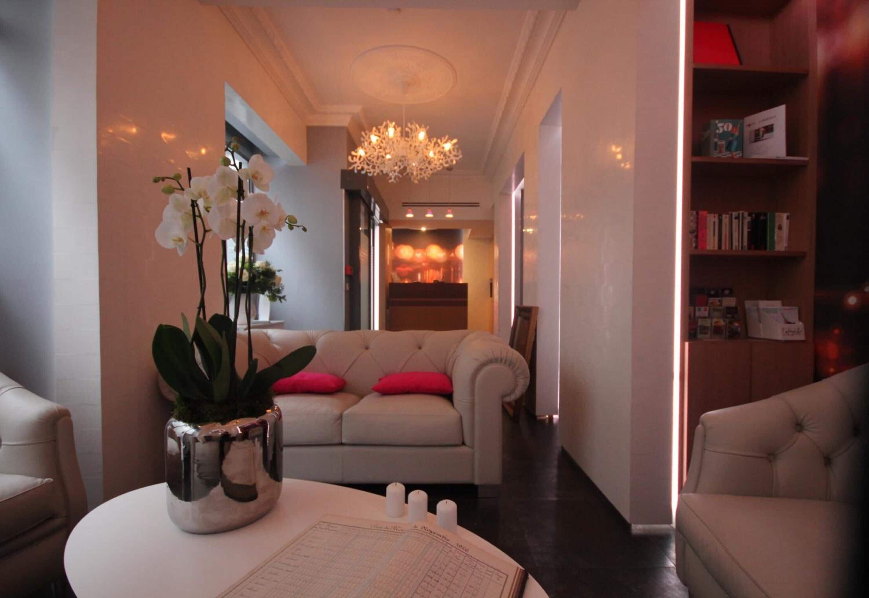 Una sala de estar moderna con un sofá, una mesa blanca con un florero de orquídeas, una araña y una estantería.
