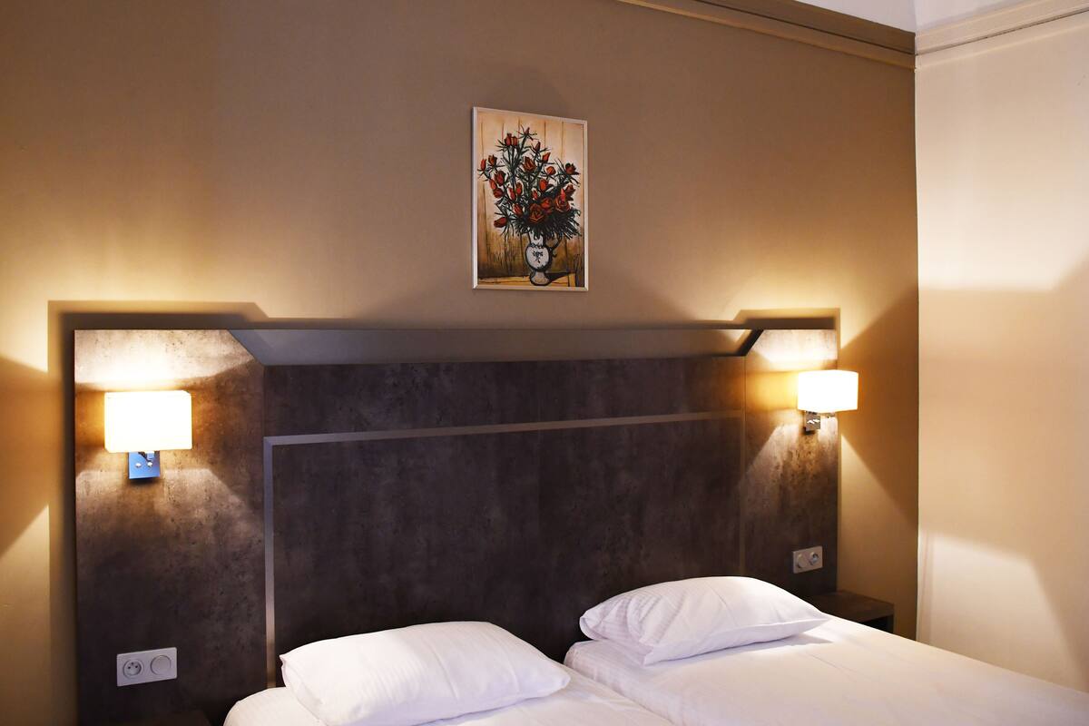 Quarto de hotel com uma cama, duas luminárias laterais, um quadro na parede e uma tomada.
