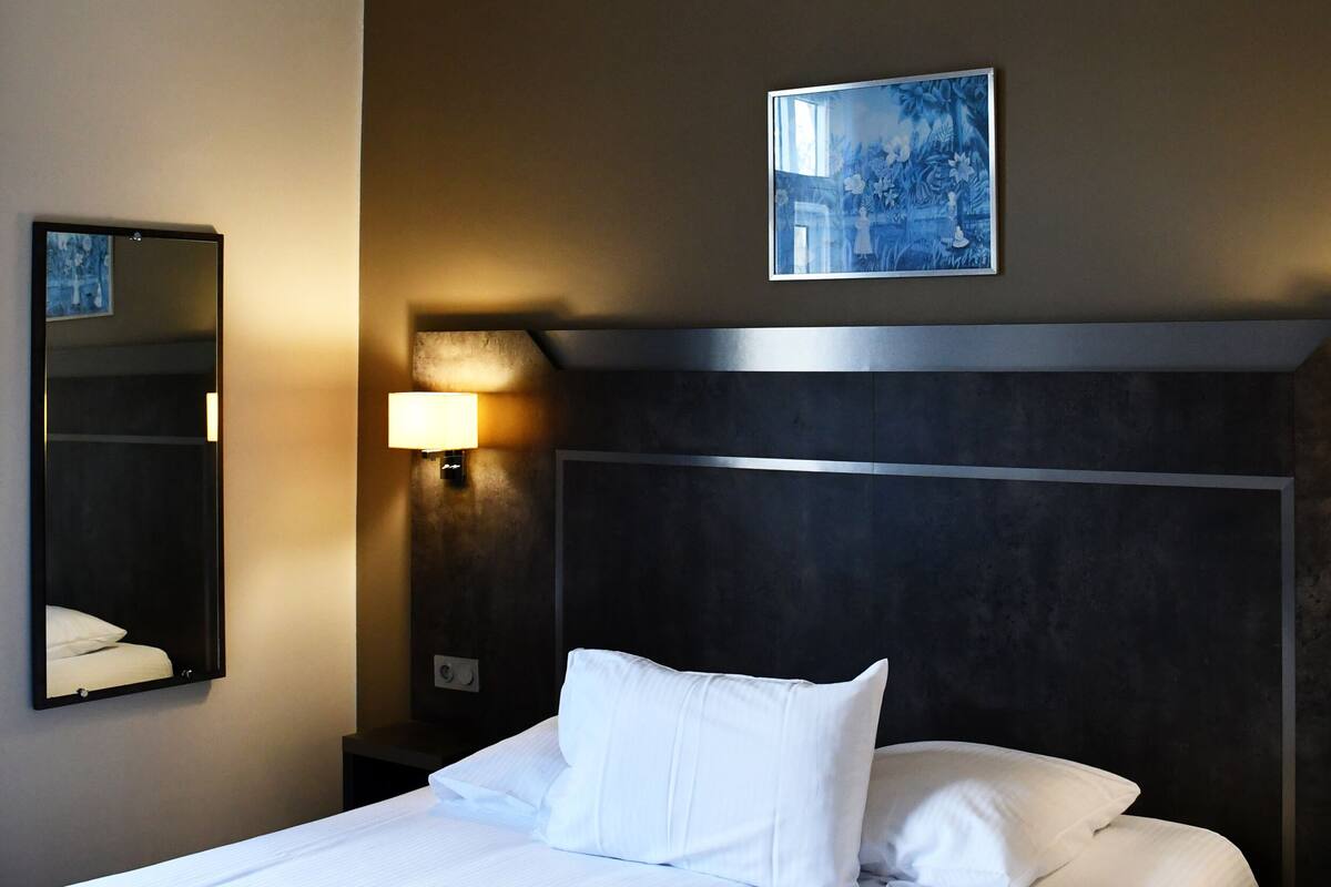 Quarto de hotel com uma cama grande, cabeceira, luminária fixada na parede, quadro emoldurado e espelho.