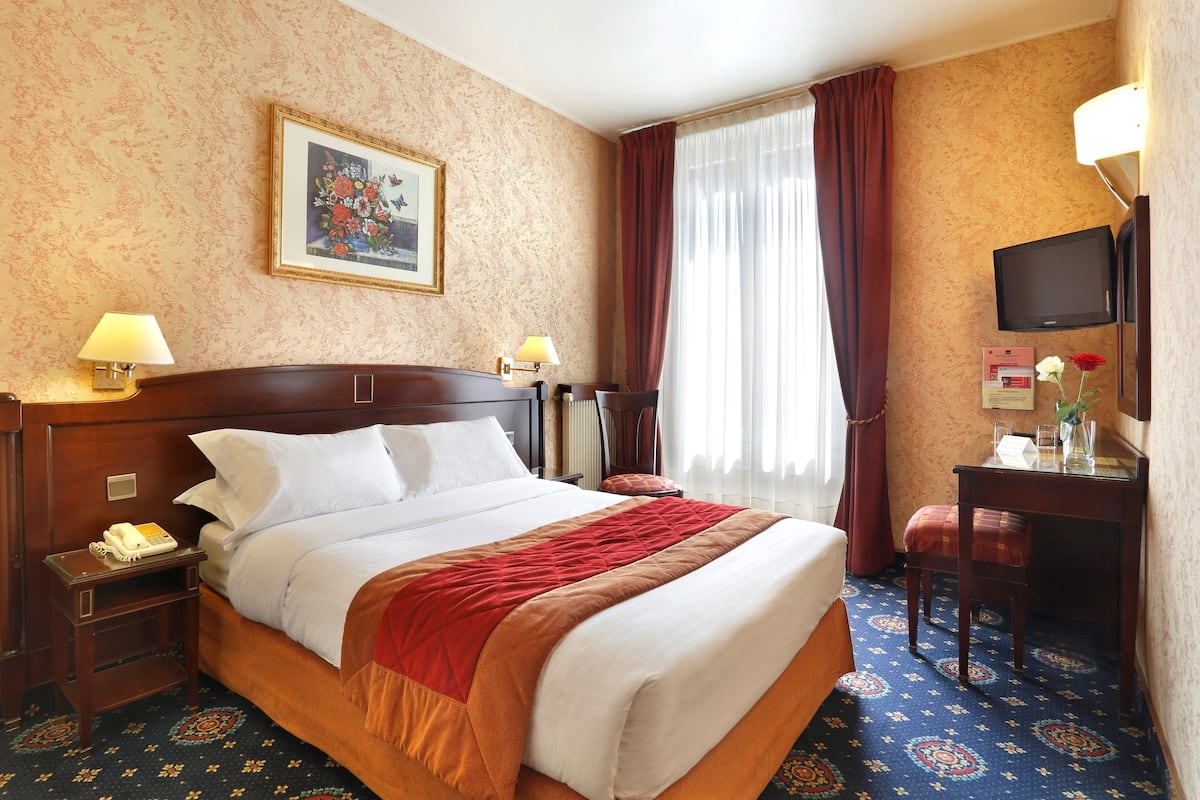 Een hotelkamer met een groot bed, twee bedlampjes, een bureau met stoel, een televisie en een raam met gordijnen.