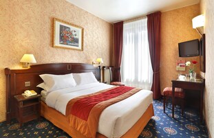 Camera d'albergo con un letto grande, due lampade ai comodini, una scrivania con una sedia, una televisione e una finestra con tende.