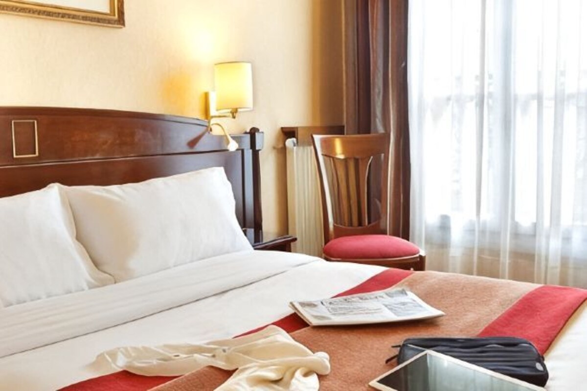 A hotel room with a bed, a chair, a lamp, and a window with curtains.