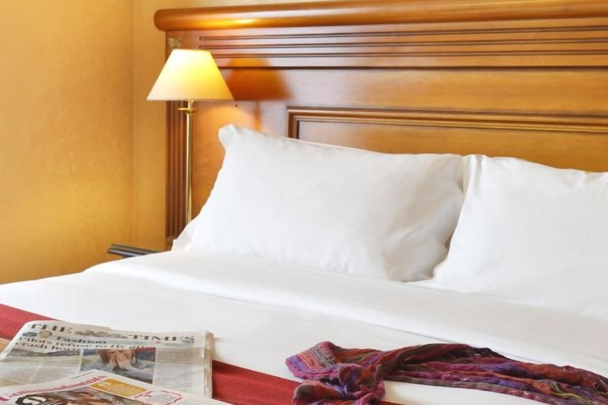 Een hotelkamer met een houten hoofdbord, wit beddengoed, een lamp en een krant op het bed.