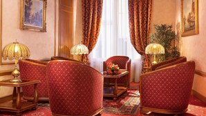 Ein Zimmer mit vier roten Polsterstühlen, zwei Lampen, einem Tisch mit Blumen und einem großen Fenster mit Vorhängen.