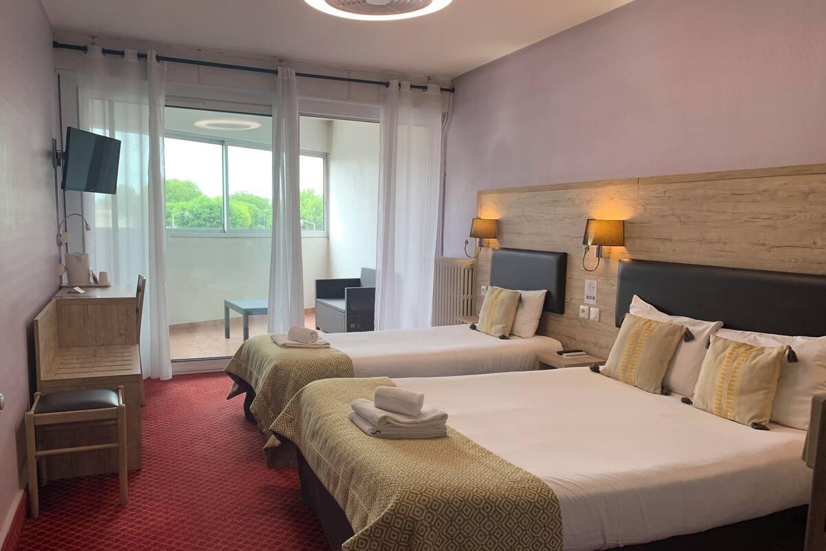 A hotel room with two beds, a desk, a chair, a TV, and a view of greenery outside.