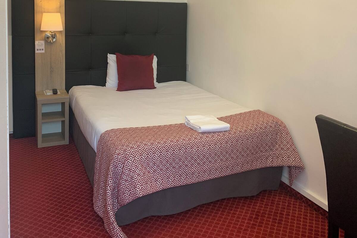 A hotel room with a bed, a bedside table with a lamp, a chair, and a unique ceiling light fixture.