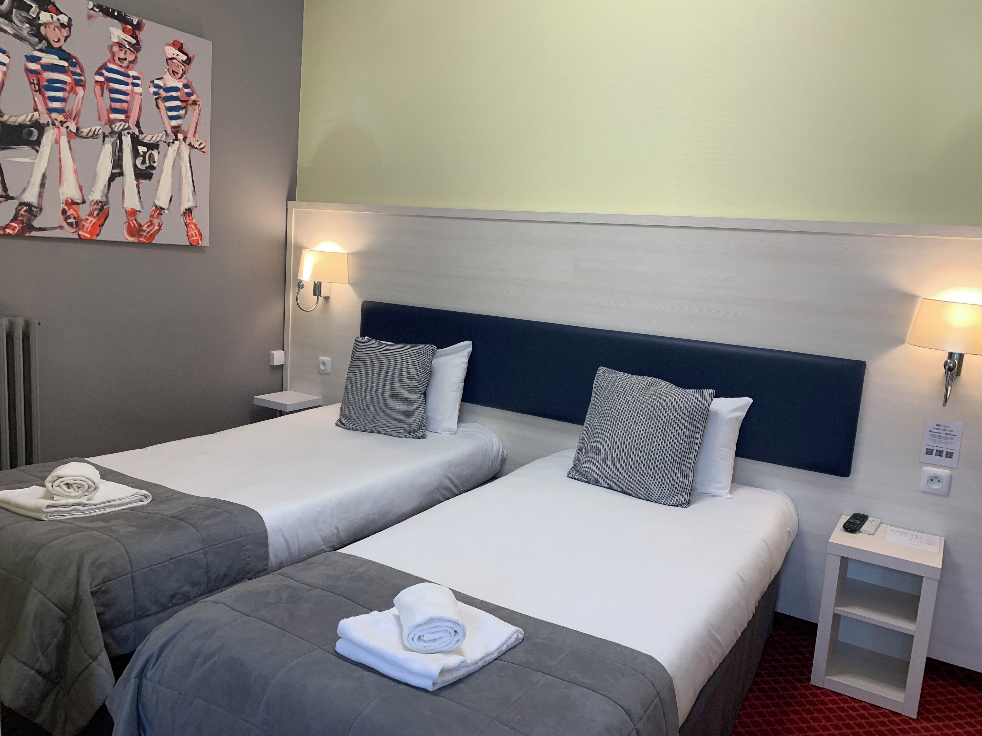 Et hotelværelse med to senge, hver med pude og en sammenrullet håndklæde. Der er et lille natbord med lampe og fjernbetjening.