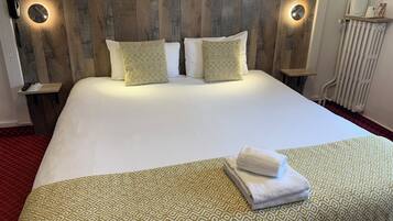 A neatly made bed with patterned pillows and a folded towel, wooden headboard with wall-mounted lights, and a small bedside table with a remote control.