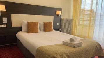 A hotel room with a large bed, two brown pillows, a patterned bedspread, a rolled towel, a nightstand with a lamp, and a window with curtains.