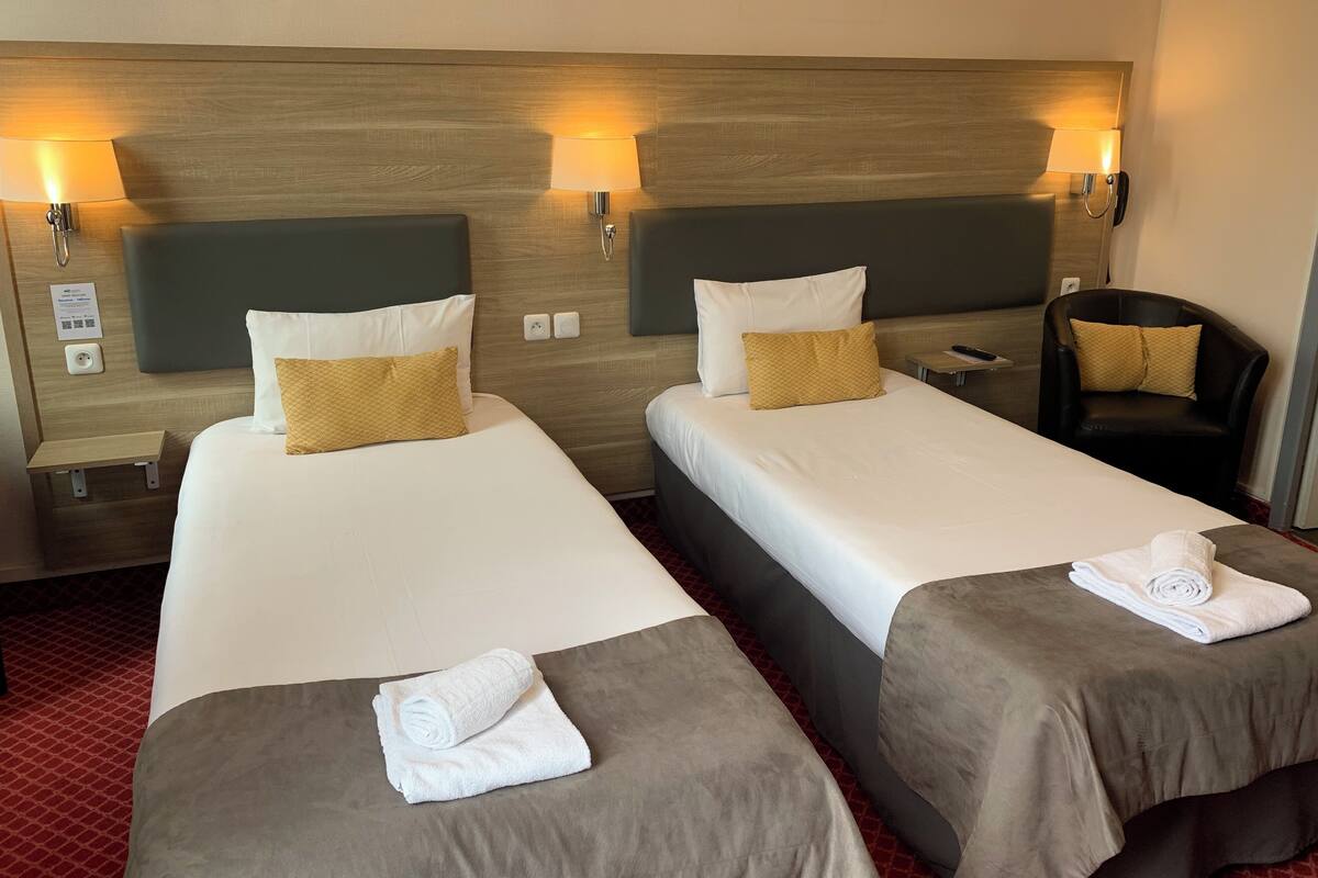 A hotel room with two beds, each with a pillow, a chair, and a small table.