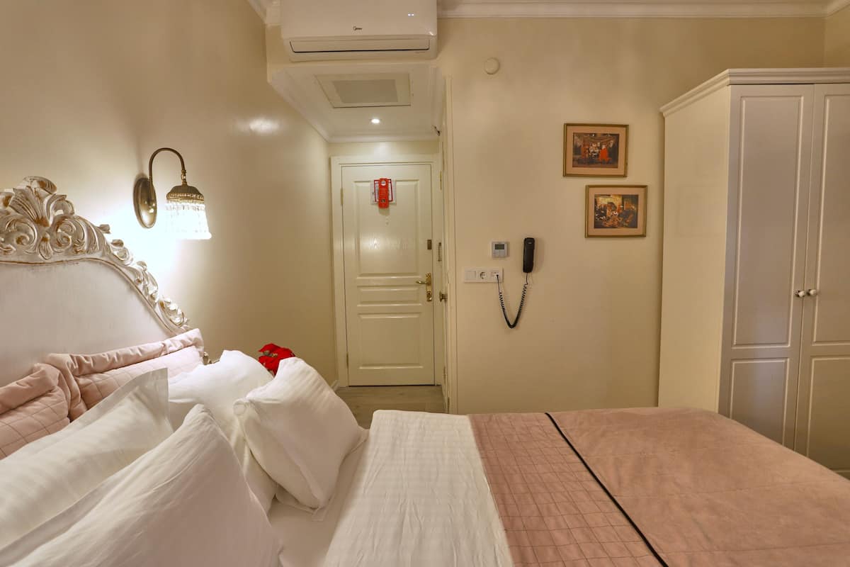 A hotel room with a bed, a bedside table, a wardrobe, a wall-mounted phone, and a door with a red emergency button.