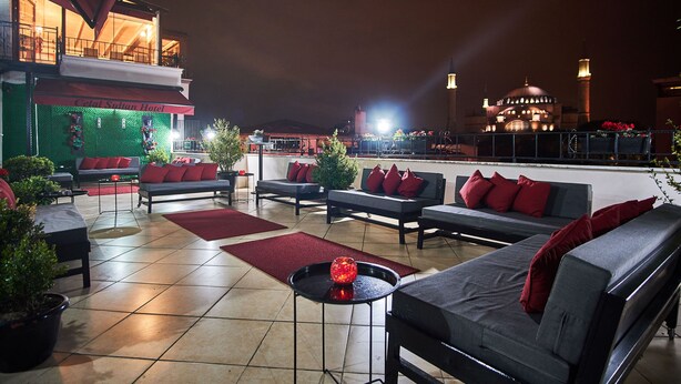 An outdoor seating area with multiple sofas and tables, a green wall with a sign, and a building with a dome and minarets in the background.