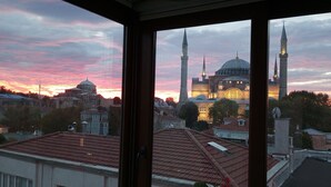 A view of a cityscape with a prominent mosque with two minarets and a dome, seen through a window with a tiled roof.