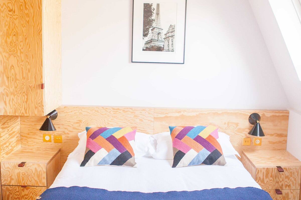 A bedroom with a wooden headboard, a bed with a blue blanket, two bedside tables with lamps, and a framed picture on the wall.
