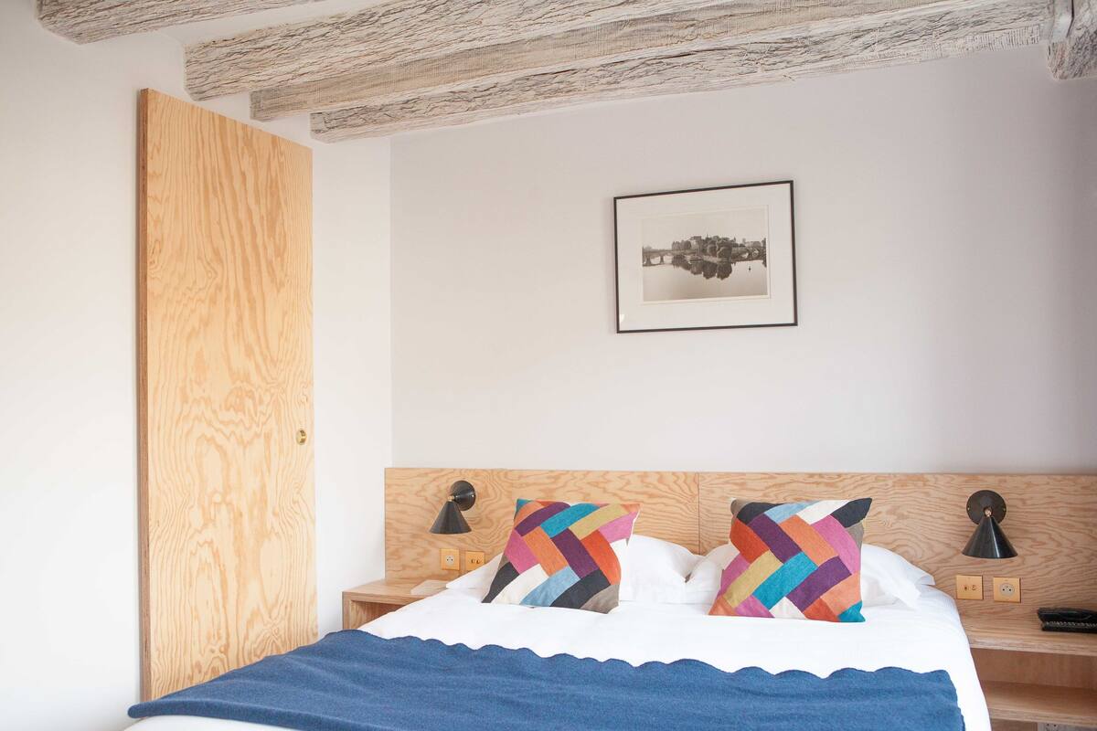 A bedroom with a wooden headboard, a bed with white linens, two colorful pillows, a framed picture on the wall, and a wooden nightstand with a lamp.